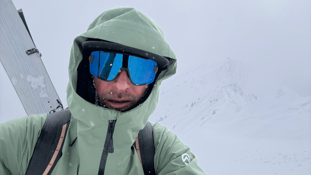 A mountain guide skier wearing a green jacket and reflective goggles stands in a snowy landscape during a snowstorm, with snowy mountains visible in the background.